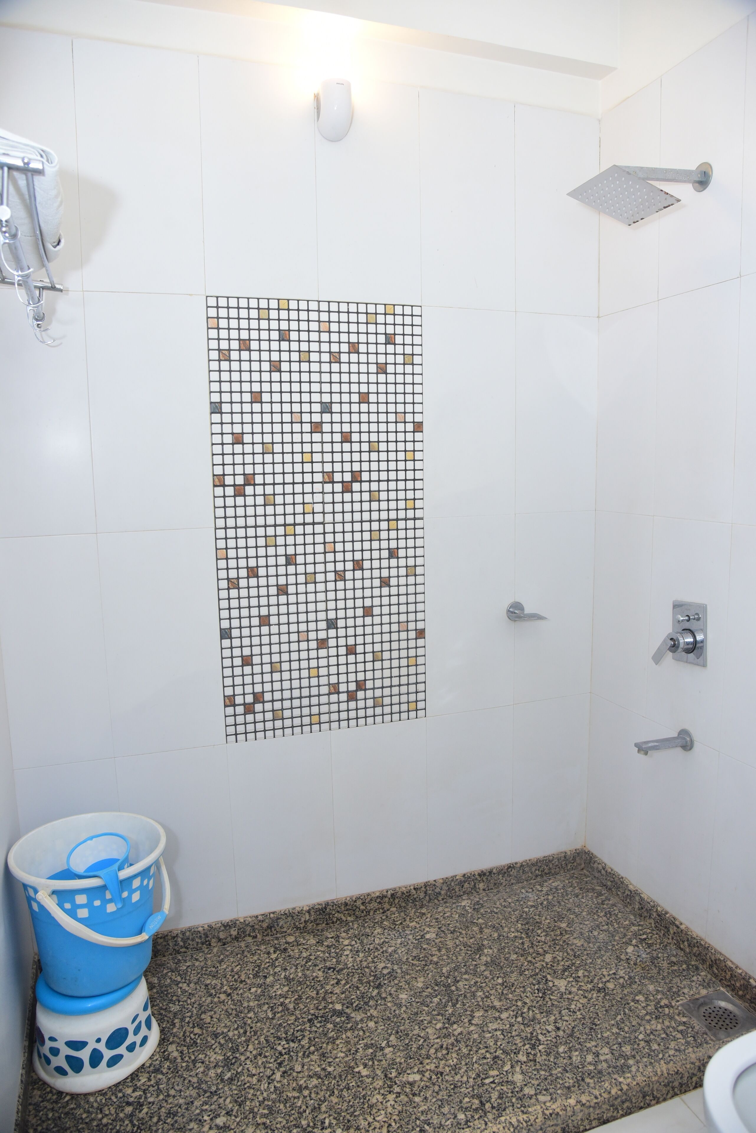 Une salle de bain avec une douche, un mur carrelé orné d’un motif en mosaïque, un sol en granit et un seau bleu et blanc.