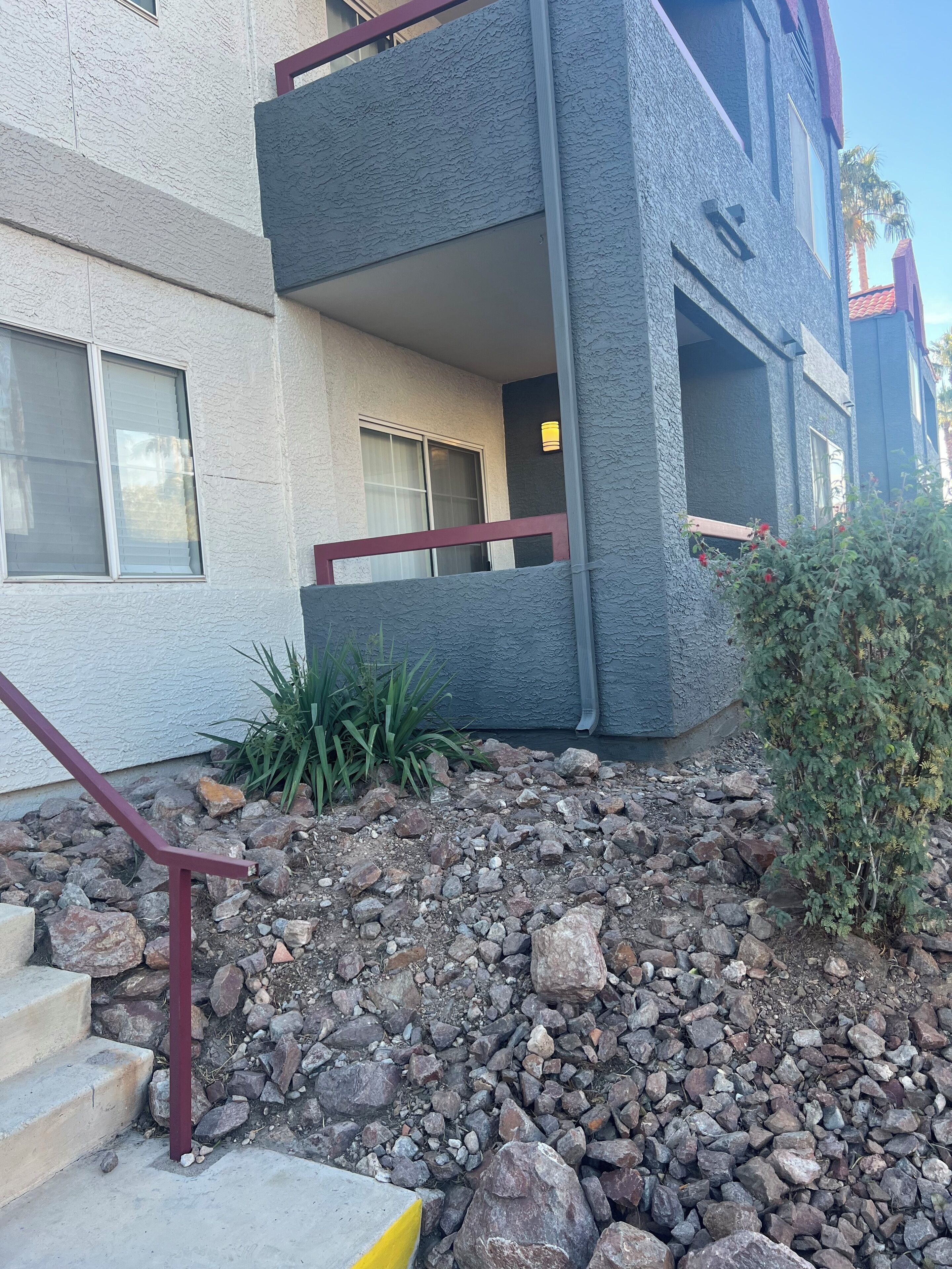 A multi-story building with a stone facade, a red railing, and a small garden area with rocks and plants.