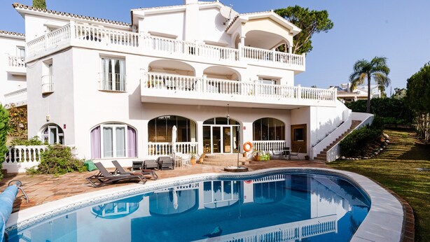 Una casa grande y blanca de dos pisos, con piscina, mobiliario exterior y una escalera que conduce a un balcón.
