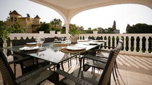Un espacio de comedor al aire libre con una mesa de vidrio y sillas de mimbre, frente a un fondo de edificios y árboles.
