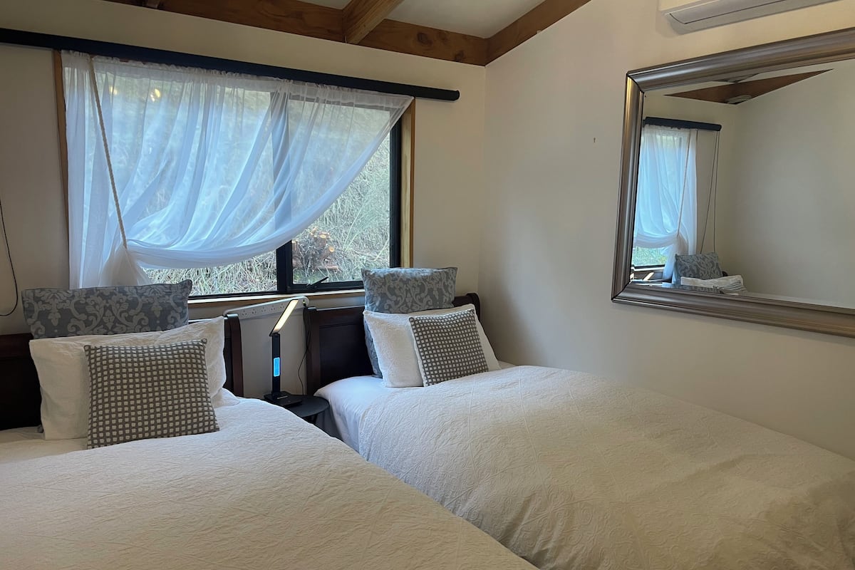 A bedroom with two beds, a bedside table, a lamp, a window with sheer curtains, and a mirror on the wall.