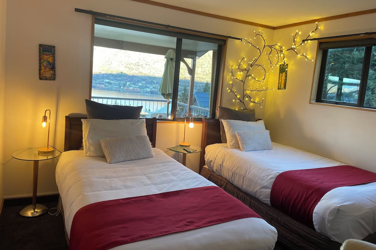 A hotel room with two beds, a large window with a view, a wall-mounted light fixture, and a small table with a lamp.