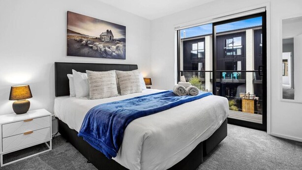 Une chambre moderne avec un grand lit, une table de chevet, une lampe de chevet, un tableau et une vue sur un immeuble par une grande fenêtre.