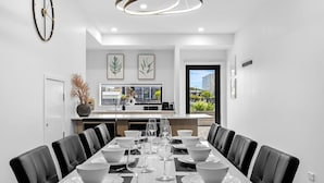 Une salle à manger moderne avec une longue table dressée pour six personnes, des chaises en cuir noir et, au fond, une cuisine avec vue.