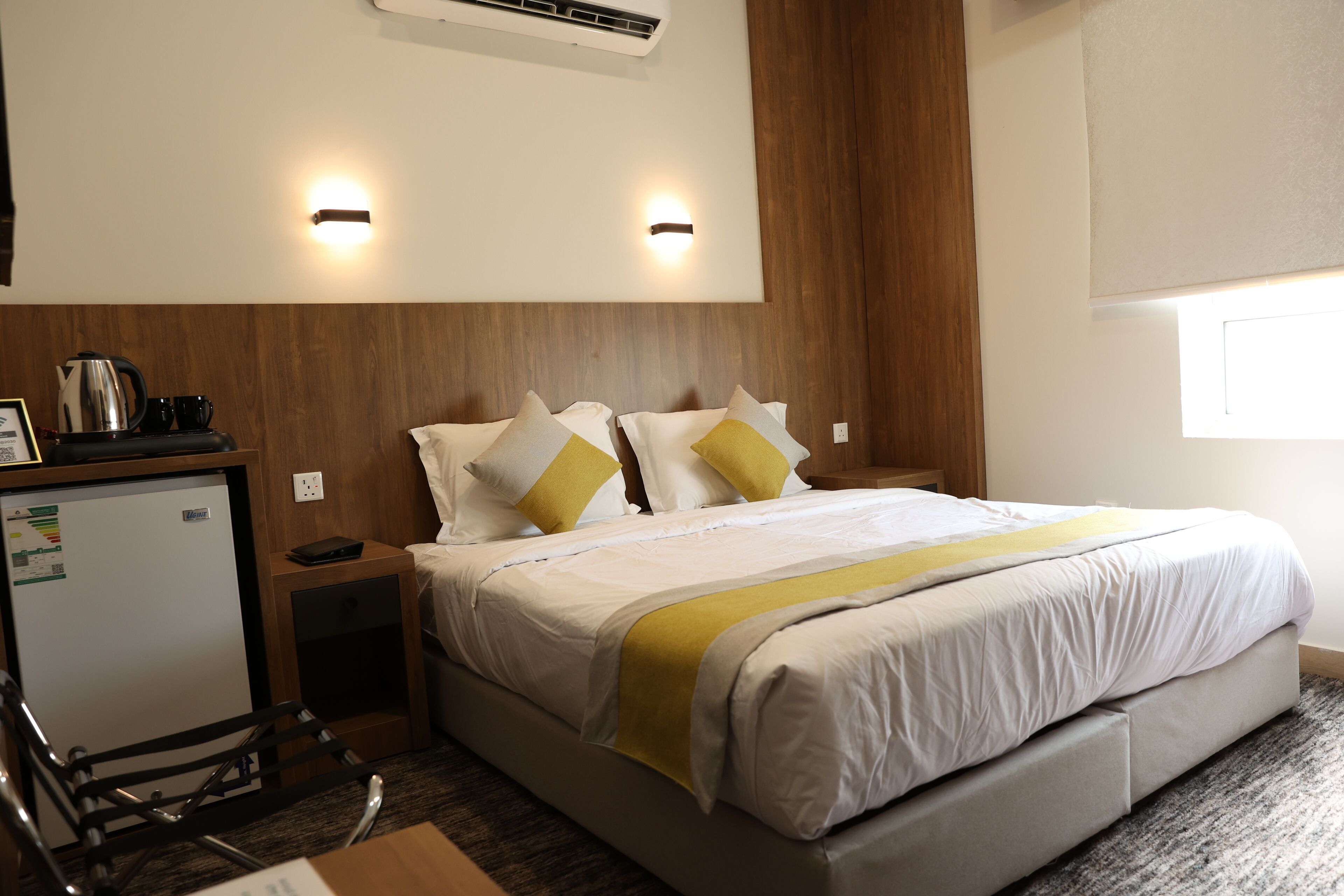 A hotel room with a bed, a desk with a kettle, a refrigerator, and a window.