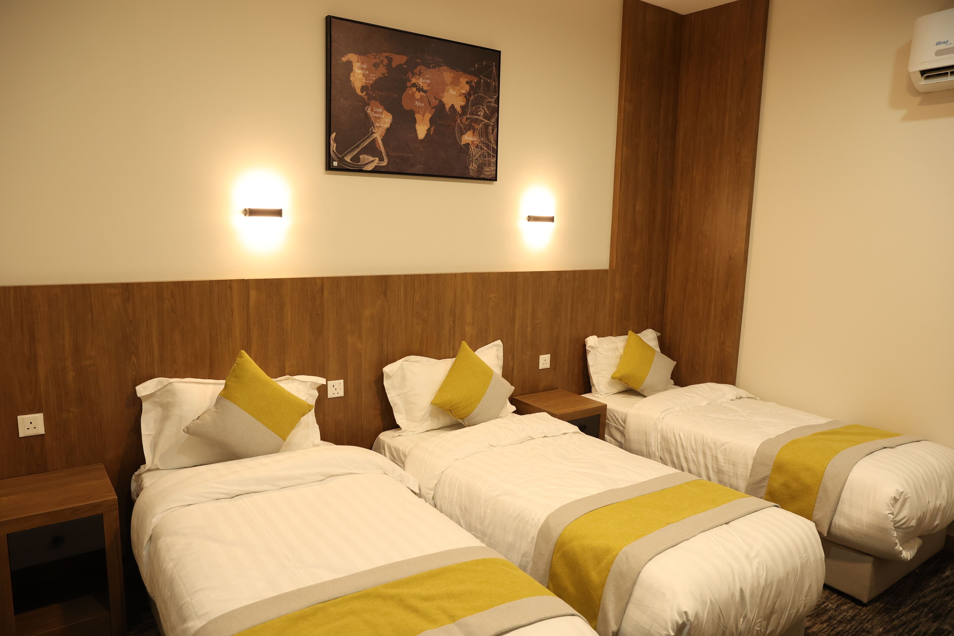 Ein Hotelzimmer mit drei Einzelbetten, jeweils mit weißer Bettwäsche und gelben Akzenten. Zwei Wandleuchten und eine Weltkarte sind an der Wand angebracht.