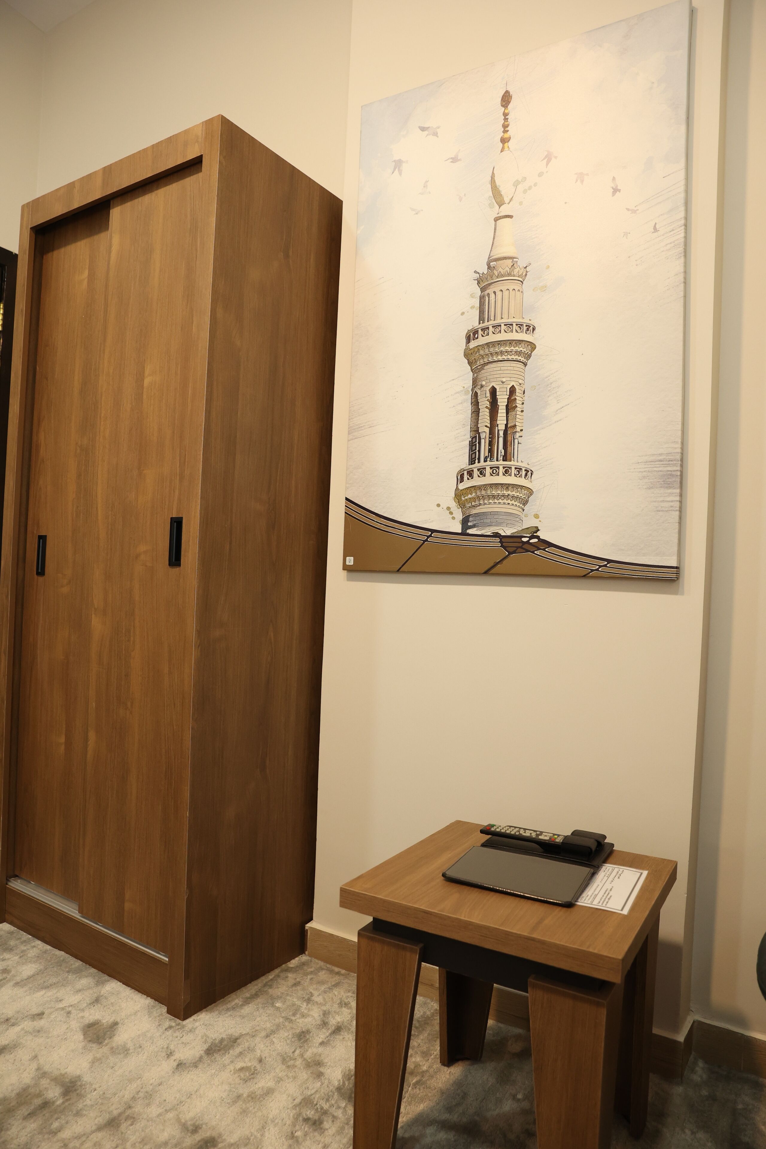 A wooden wardrobe, a small wooden table with a black notebook, and a framed picture of a minaret.
