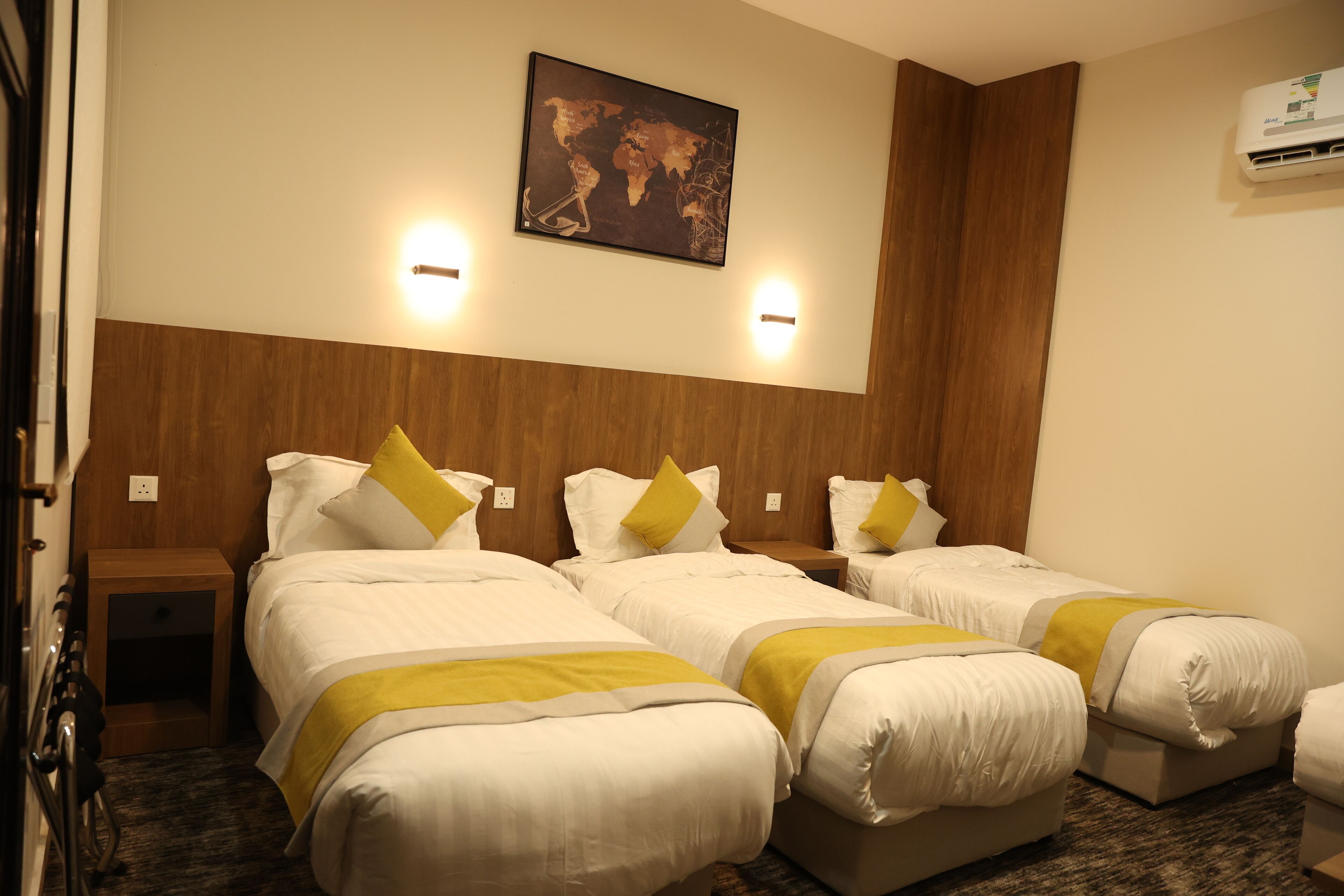 A hotel room with three single beds, each with white bedding and a yellow accent. There is a wooden headboard, a world map on the wall, and a wall-mounted air conditioner.