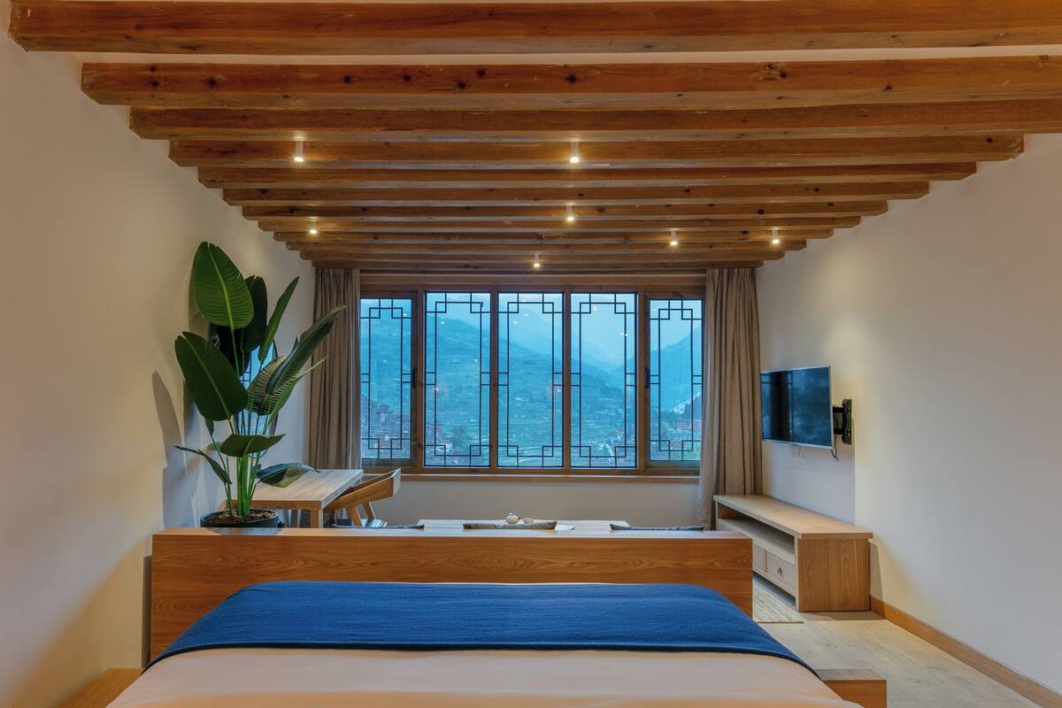 Quarto com uma cama grande, teto de madeira, janela com vista para montanhas, uma planta e uma TV na parede.