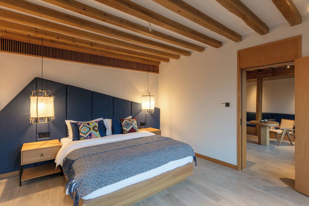 Quarto moderno com teto de madeira, cama grande com roupa de cama estampada, luminárias de cabeceira e vista para uma sala de jantar.