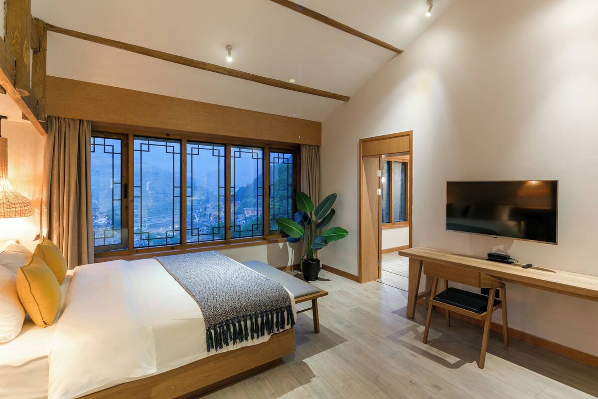 Quarto de hotel moderno com uma cama grande, uma escrivaninha, uma TV e vista para o horizonte urbano pelas janelas amplas.
