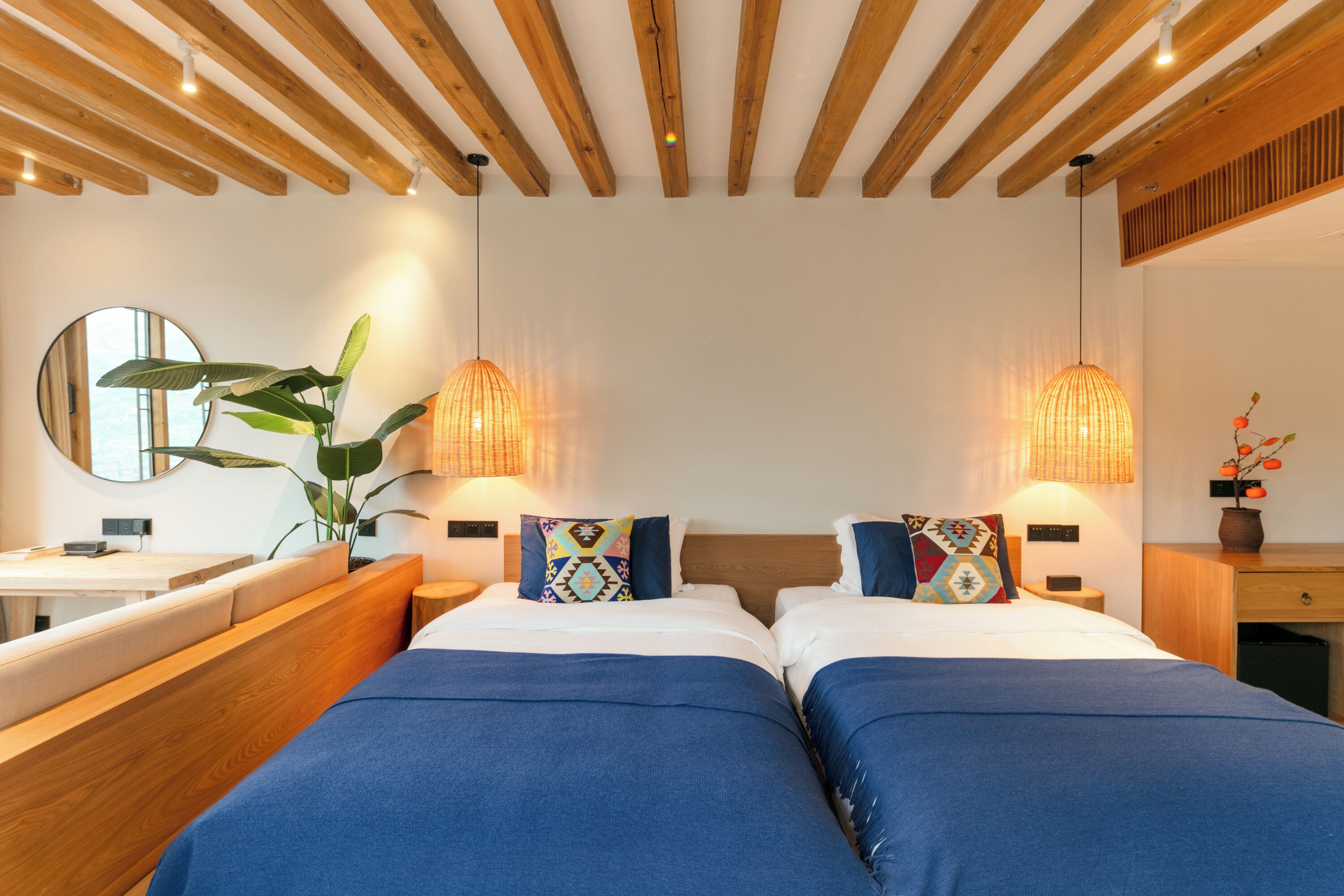 A modern hotel room with a wooden ceiling, two beds with blue bedding, a round mirror, a potted plant, and two wall-mounted lamps.