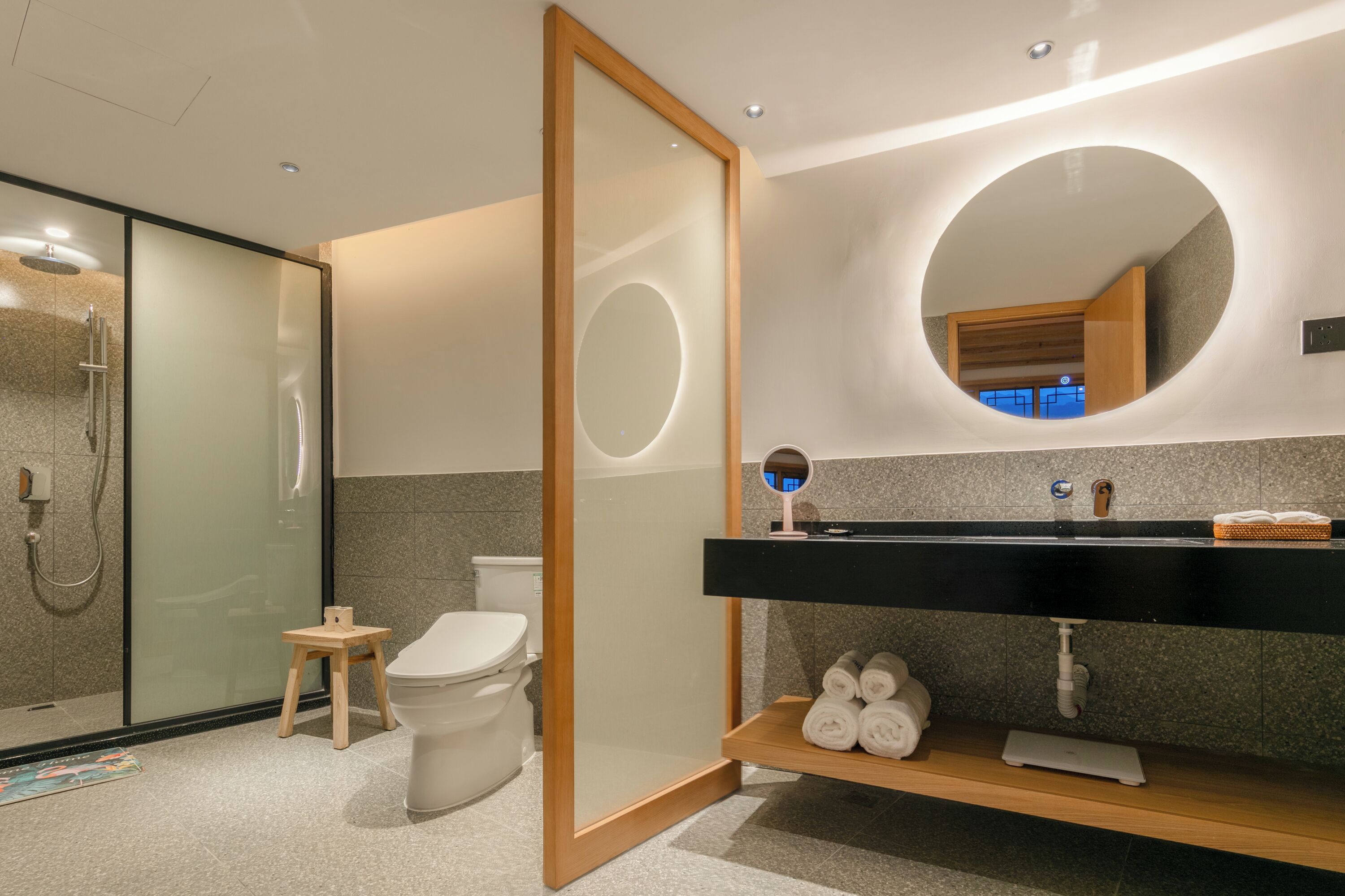 A modern bathroom with a glass-enclosed shower, a wooden stool, a toilet, a black vanity with a mirror, and a towel rack.