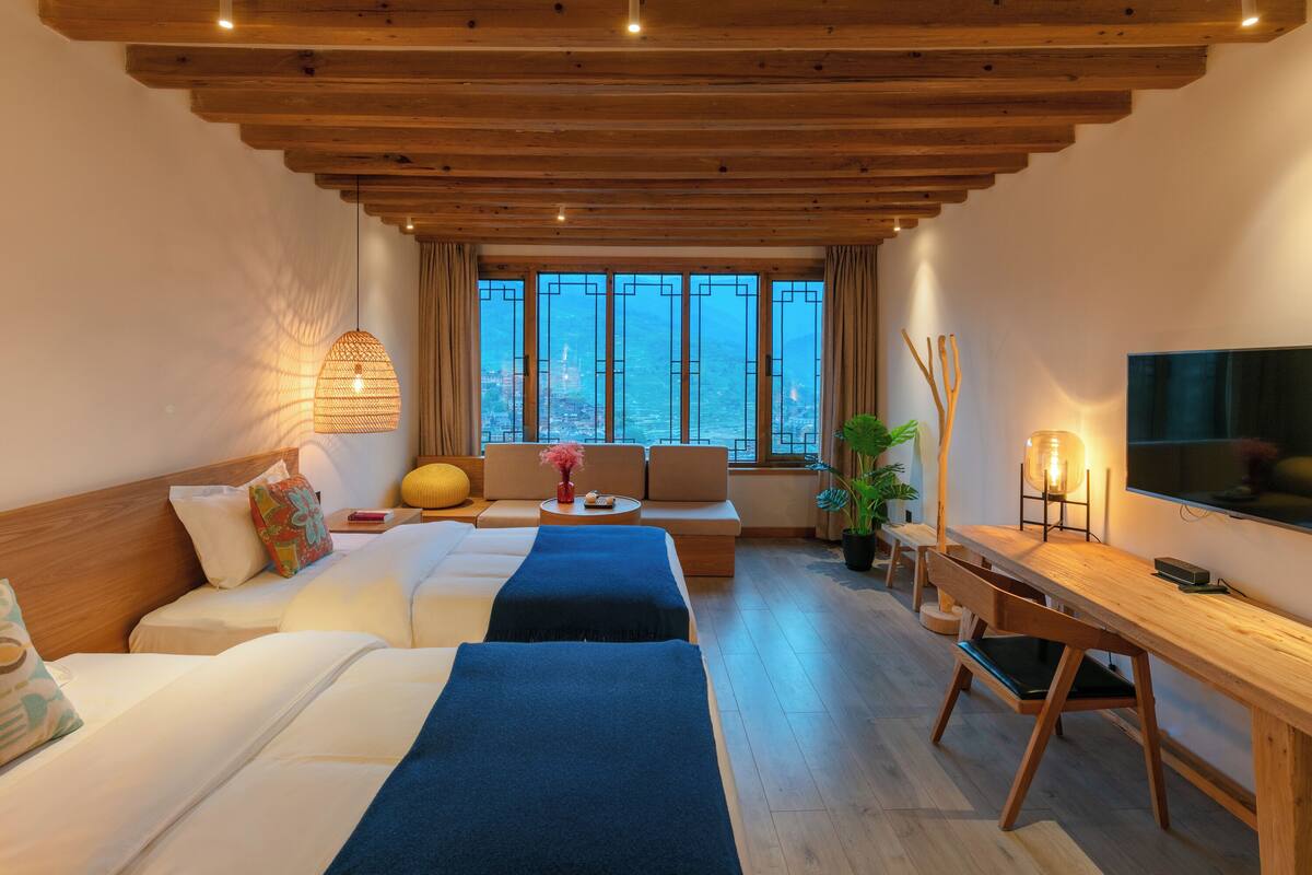 Quarto de hotel com teto de madeira, uma janela grande com vista para a cidade, duas camas, um sofá, uma escrivaninha e uma televisão.