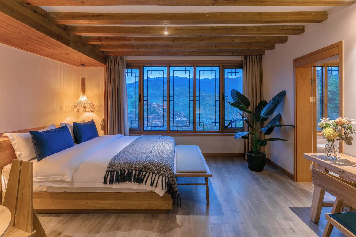 Quarto com cama grande, vigas de madeira, vista para montanhas, planta em vaso e um banco de madeira.