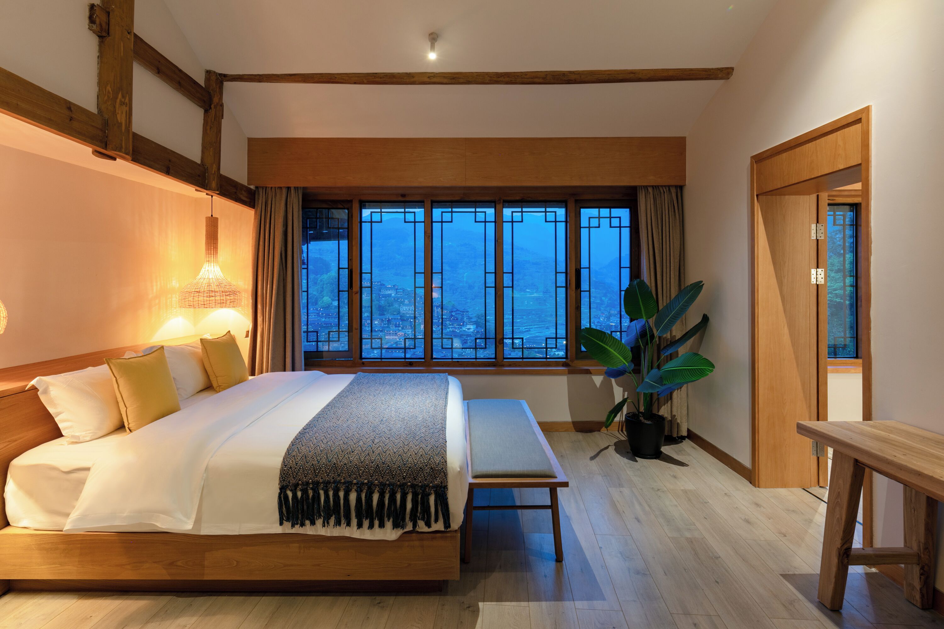 Un dormitorio amplio con una cama grande, lámparas de noche, un banco de madera y vistas a la ciudad a través de ventanas amplias.