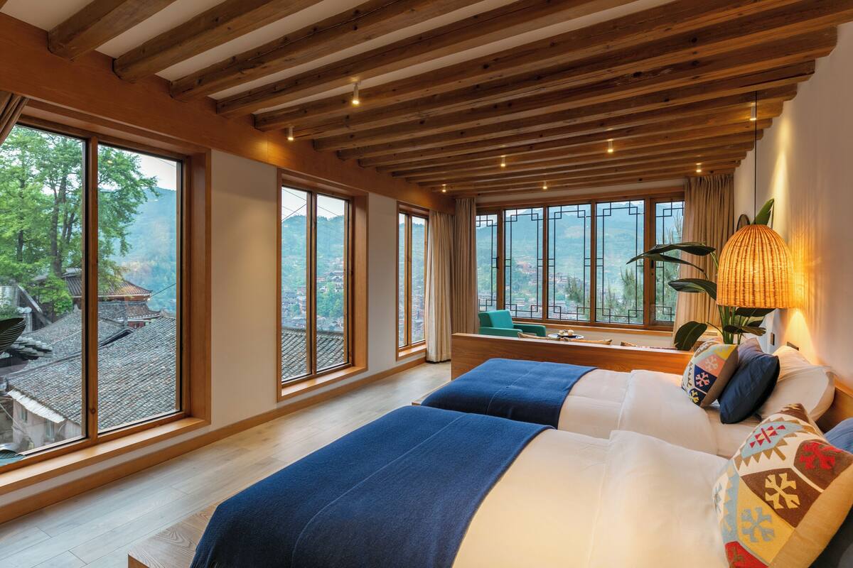 Quarto de hotel com grandes janelas, uma cama com roupa de cama azul e branca, uma escrivaninha e um abajur.