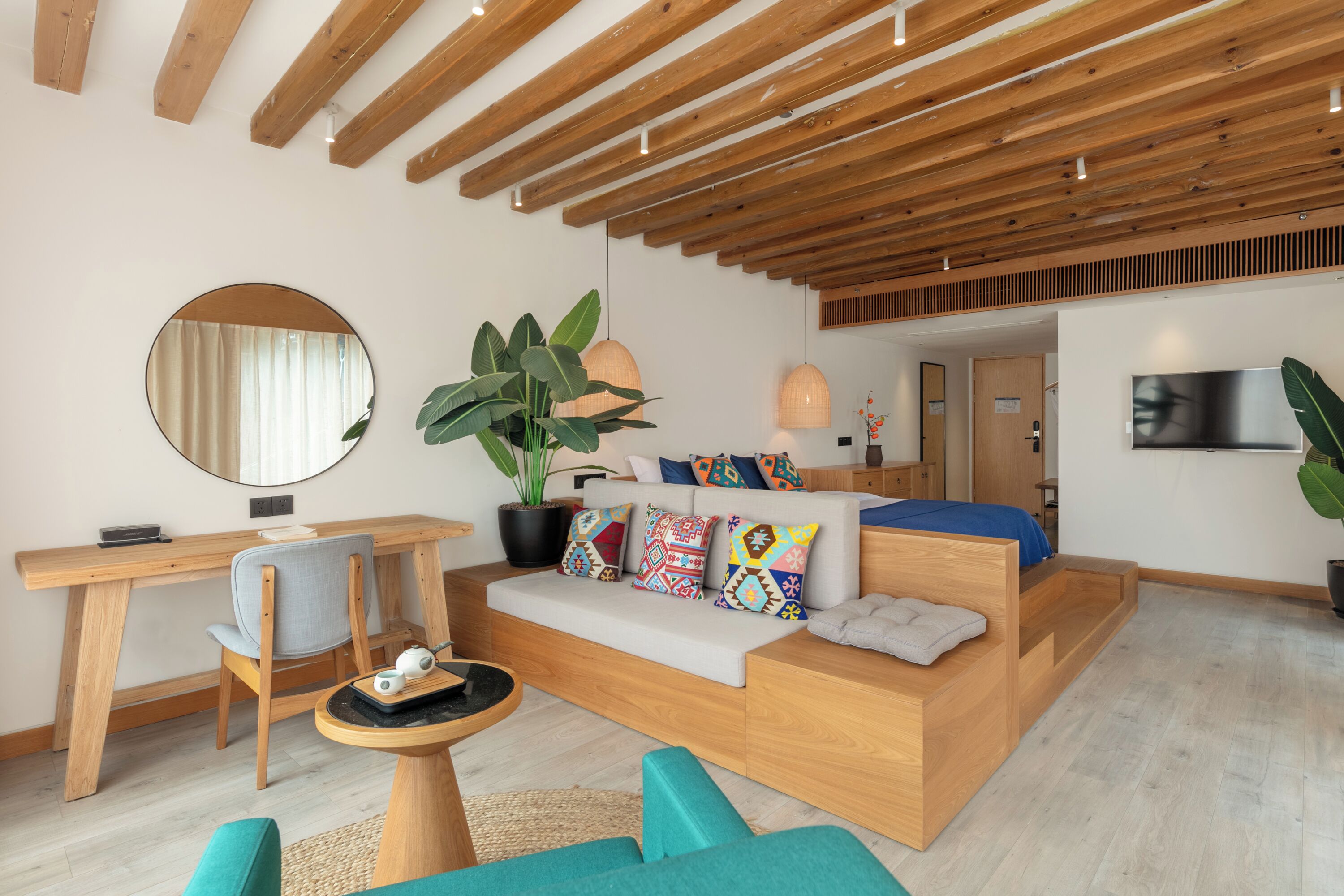 Un salon moderne avec un plafond en bois, un grand miroir rond, un canapé agrémenté de coussins colorés, un bureau et une grande plante.