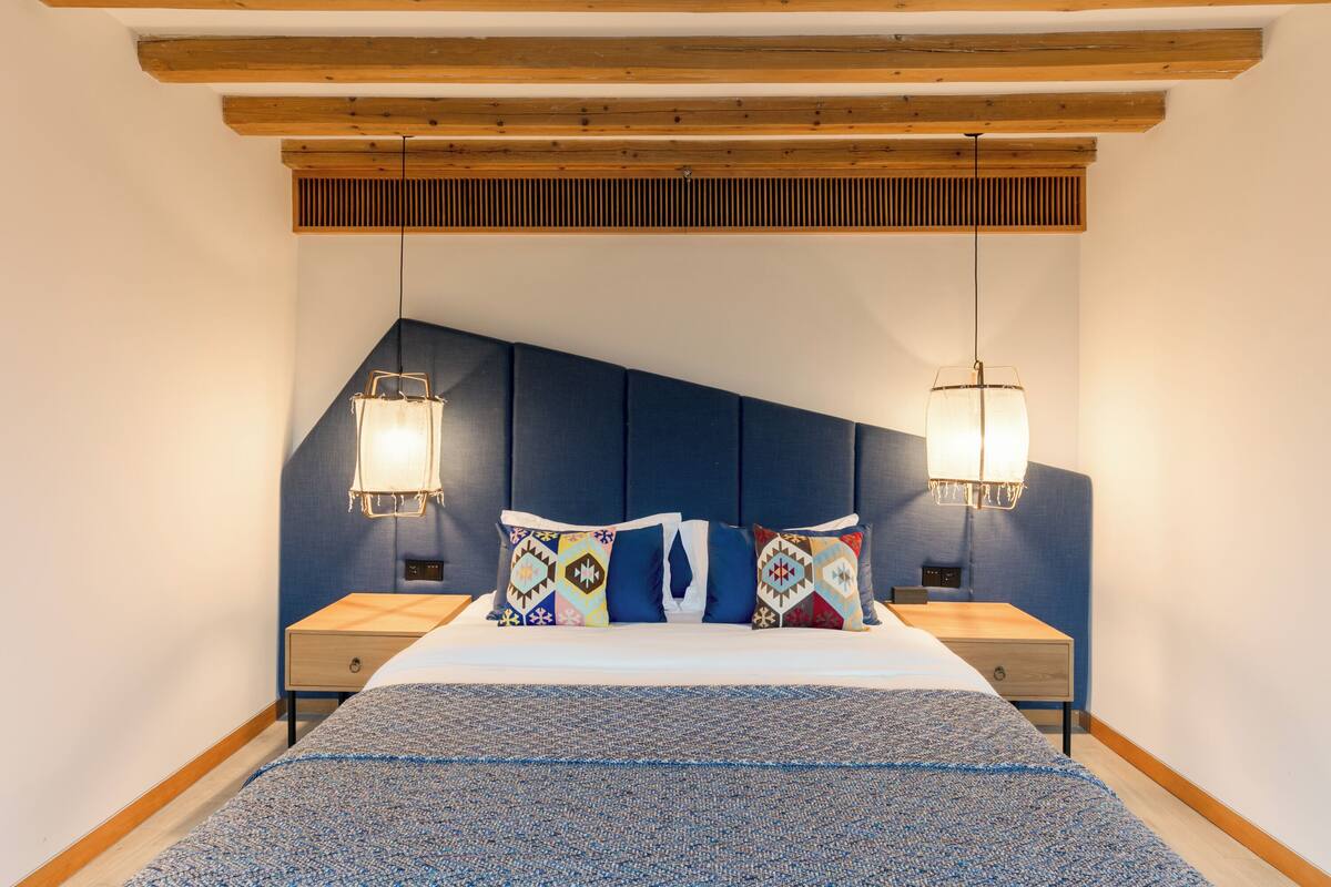 Um quarto com teto inclinado, cama com almofadas estampadas, duas mesas de cabeceira e luminárias pendentes.