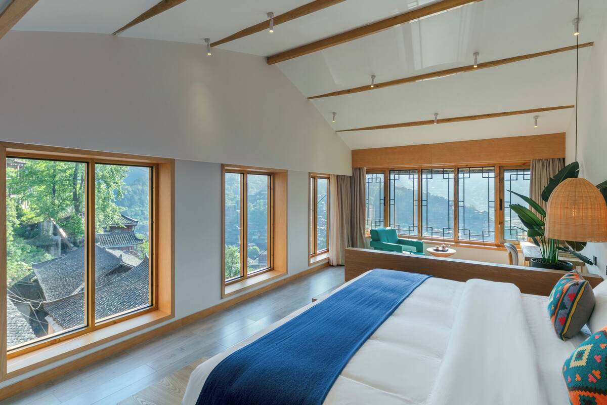 Quarto espaçoso com grandes janelas, uma cama, uma escrivaninha e vista para um edifício tradicional.