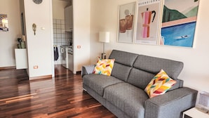 A modern living room with a grey sofa, colorful cushions, wooden flooring, a kitchen in the background, and artwork on the wall.