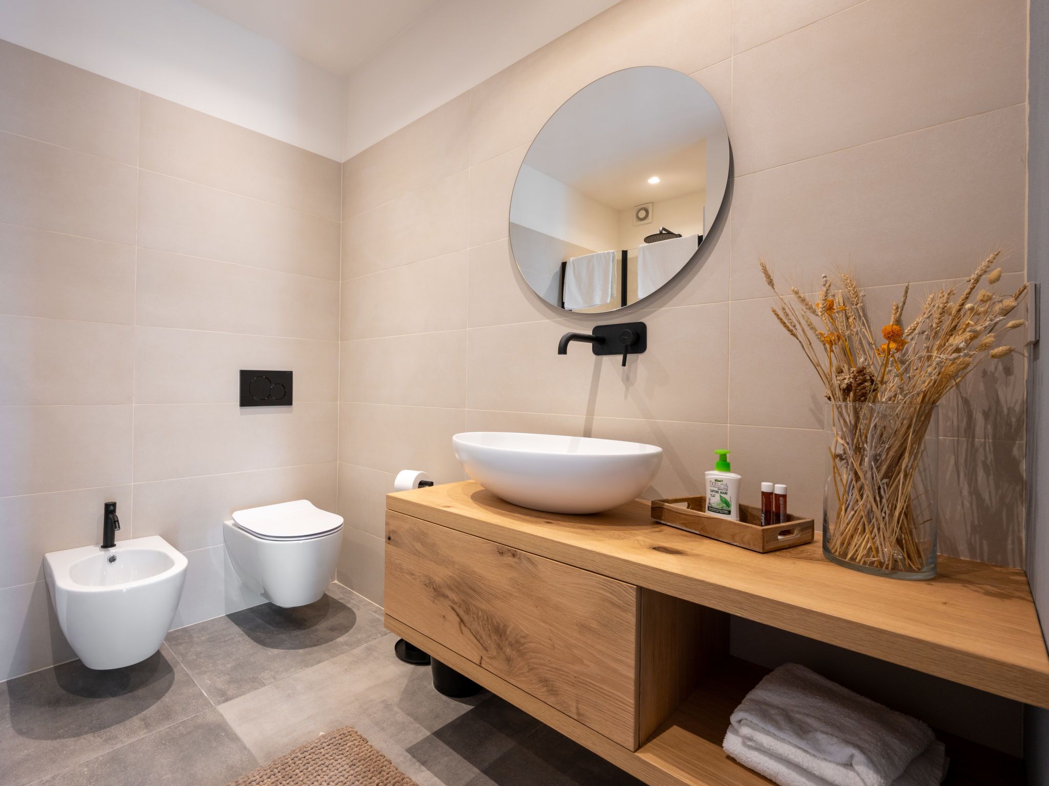 A modern bathroom with a large round mirror, a vessel sink, a bidet, and a wooden vanity.