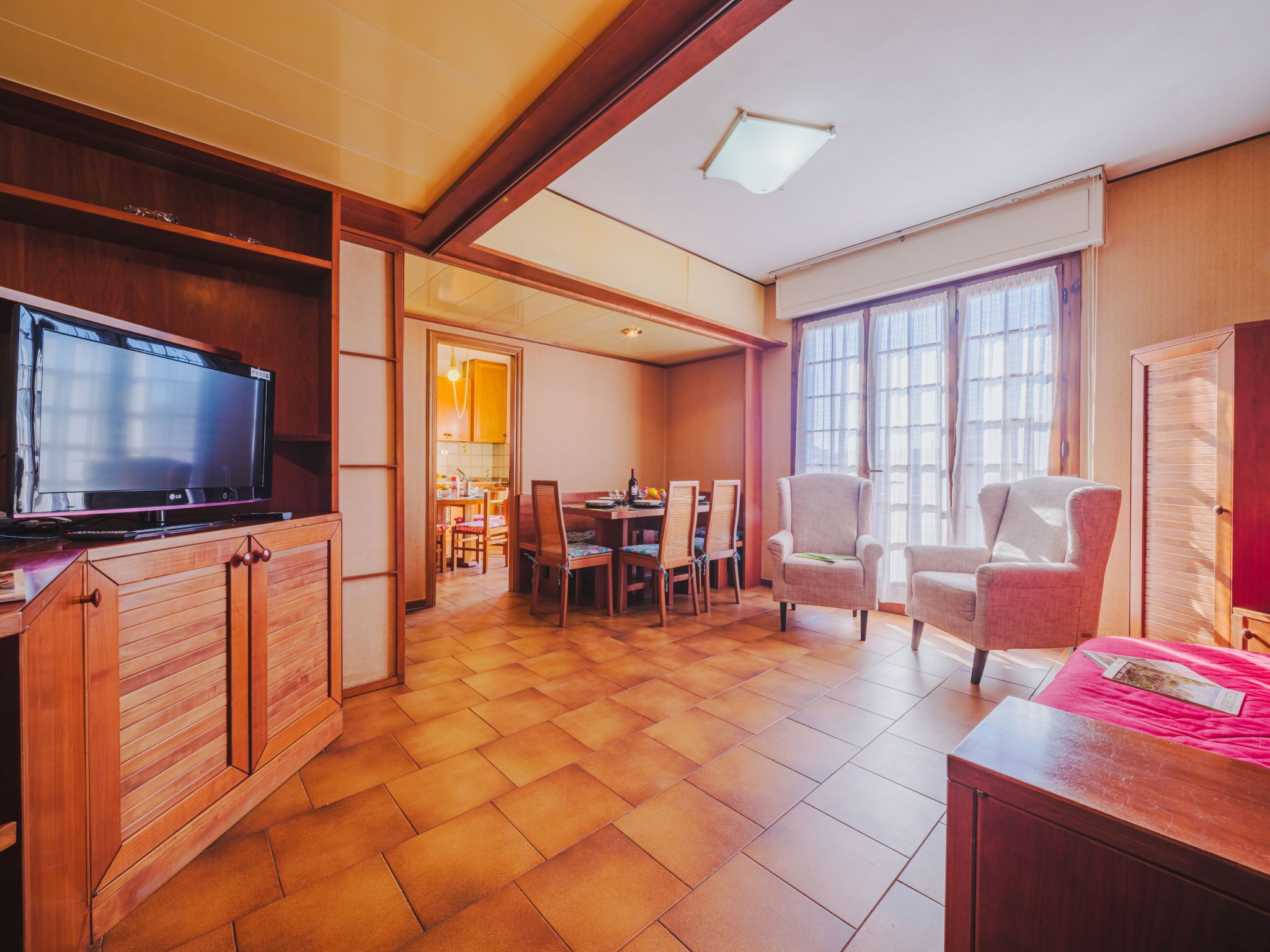 A spacious living area with a television, wooden furniture, a dining table, and large windows.
