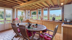 A wooden interior with a round table and chairs, large windows, and a kitchen area. - 2 Chaumières normande by Interhome (Le perrey)