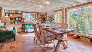 A cozy room with a wooden dining table, wicker chairs, a green sofa, and bookshelves filled with books. - 2 Chaumières normande by Interhome (Le perrey)