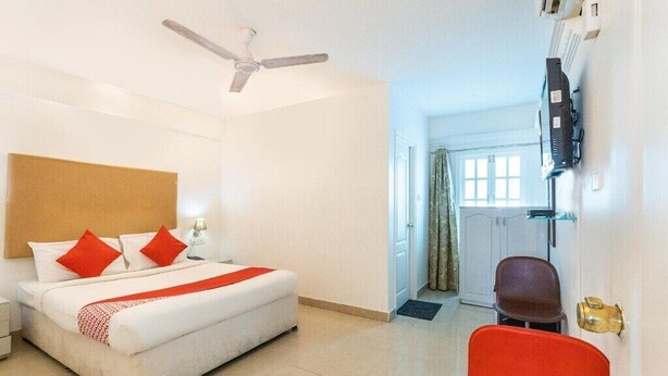 A hotel room with a bed, a ceiling fan, a television mounted on the wall, and a door leading to another room. - Hotel Fiesta Grand (Bengaluru)