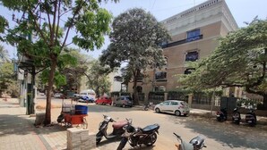 A paved area with parked motorcycles and a few cars, surrounded by trees and a building with multiple windows. - Hotel Fiesta Grand (Bengaluru)