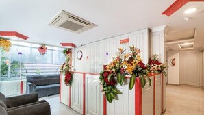 A reception area with a white reception desk, red accents, and floral decorations. There are grey sofas and a large window providing a view of the outdoors. - Hotel Fiesta Grand (Bengaluru)