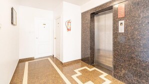 A hallway with a marble floor, a white door, and an elevator with a security sign. - Hotel Fiesta Grand (Bengaluru)