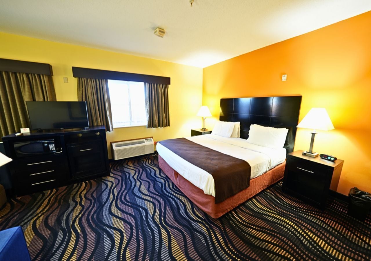 Quarto de hotel com cama, televisão, escrivaninha e mesa de cabeceira.