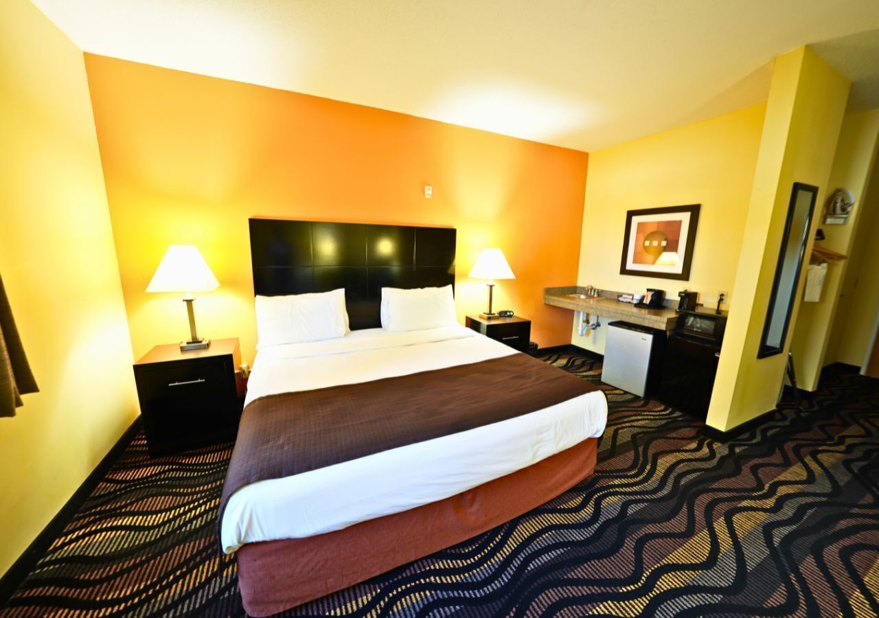 Een hotelkamer met een groot bed, twee nachtkastjes met lampen, een klein kitchenette en een ingelijst schilderij aan de muur.