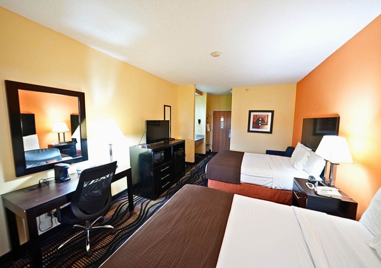 Habitación de hotel con dos camas, un escritorio con computadora, un televisor y un espejo.