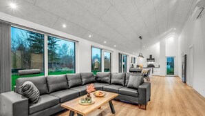 Un salon spacieux avec un grand canapé en cuir noir, une table basse en bois et une vue sur un jardin à travers de grandes fenêtres.