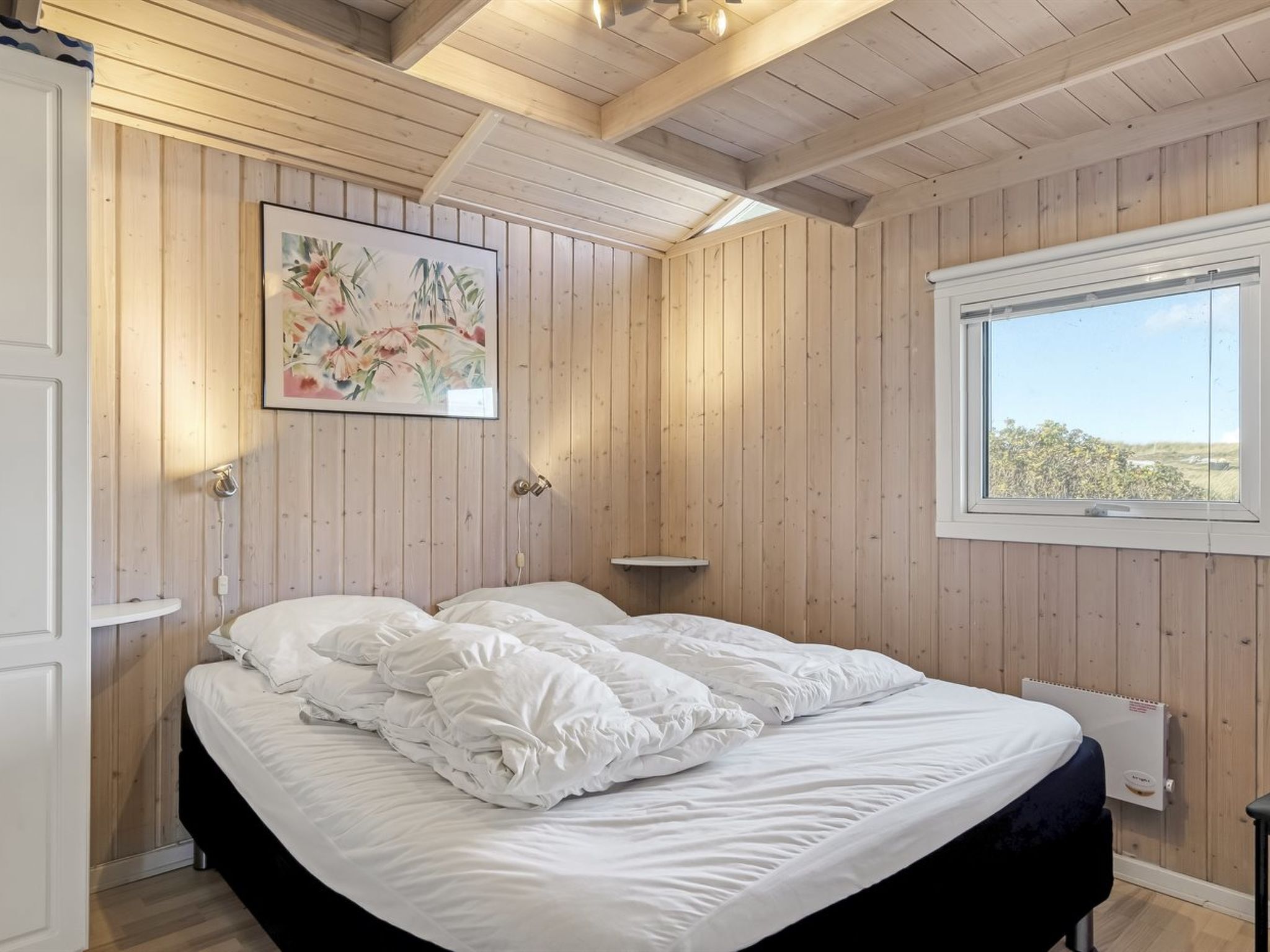 Un dormitorio con techo de madera, una cama con ropa de cama blanca, una ventana con vistas y un radiador fijado a la pared.