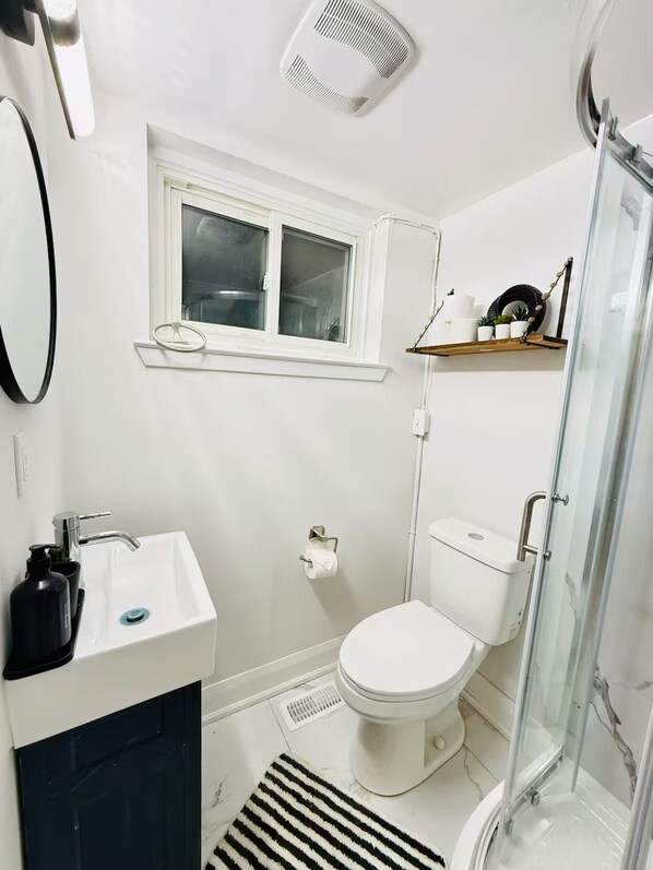 A bathroom with a white sink, toilet, and shower. There is a window above the sink and a shelf with toiletries. - Pet friendly private backyard Garden studio (Pickering)