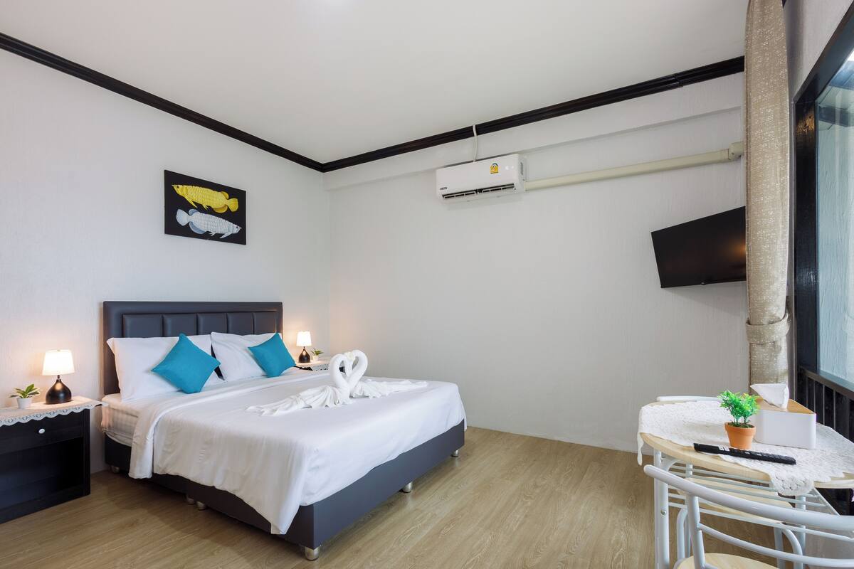 A hotel room with a bed, bedside table, lamp, air conditioning unit, and a small table with a plant.