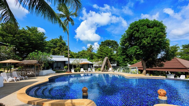 A swimming pool with a tiled edge, surrounded by lounge chairs and umbrellas, with trees and a building in the background. - Peace Garden Resort Koh Samui (Koh Samui)