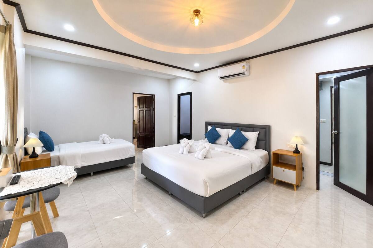 A modern hotel room with a large bed, two smaller beds, a desk with a chair, and a small table with a lamp.