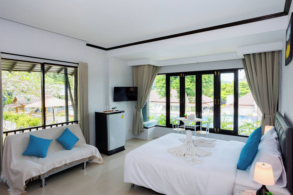 A hotel room with a bed, a sofa, a TV, a desk, and a view of greenery outside.
