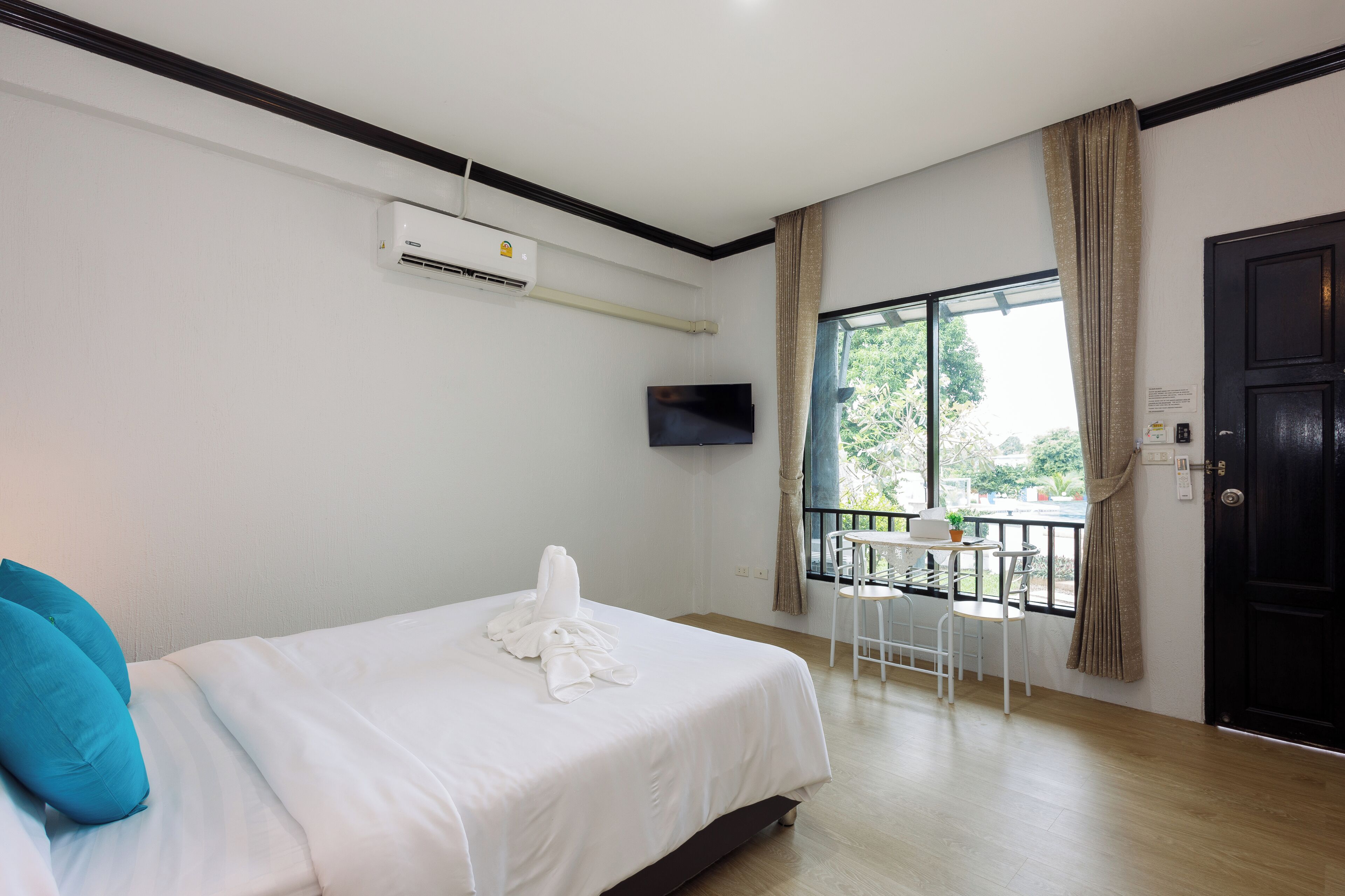 A hotel room with a bed, a TV mounted on the wall, a window with curtains, a small table with a chair, and a door.