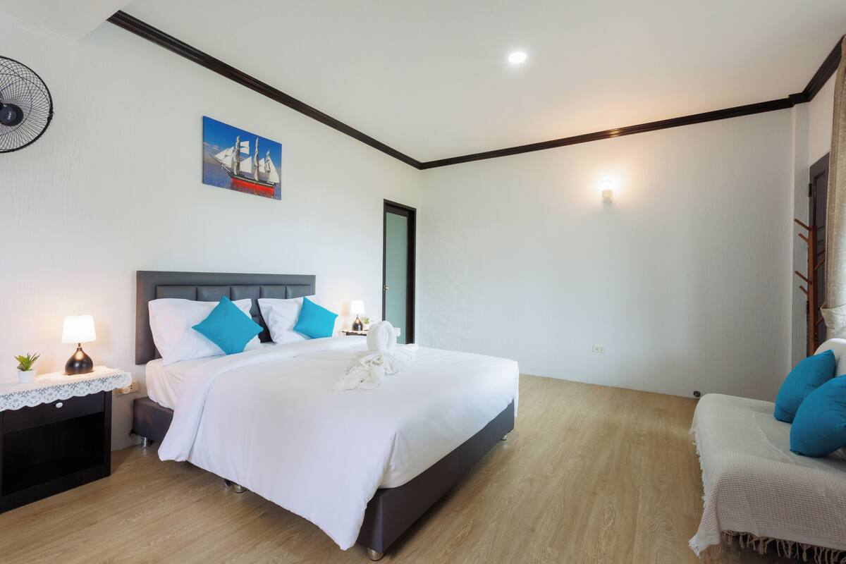 A modern hotel room with a large bed, bedside tables, a nightstand, a wall-mounted fan, a painting, and a door.