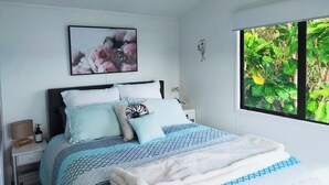 Een slaapkamer met een bed, nachtkastje, een aan de muur bevestigde televisie en een raam met uitzicht op groen.