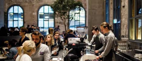 A busy cafe with a rustic brick wall, large windows, and a coffee machine.