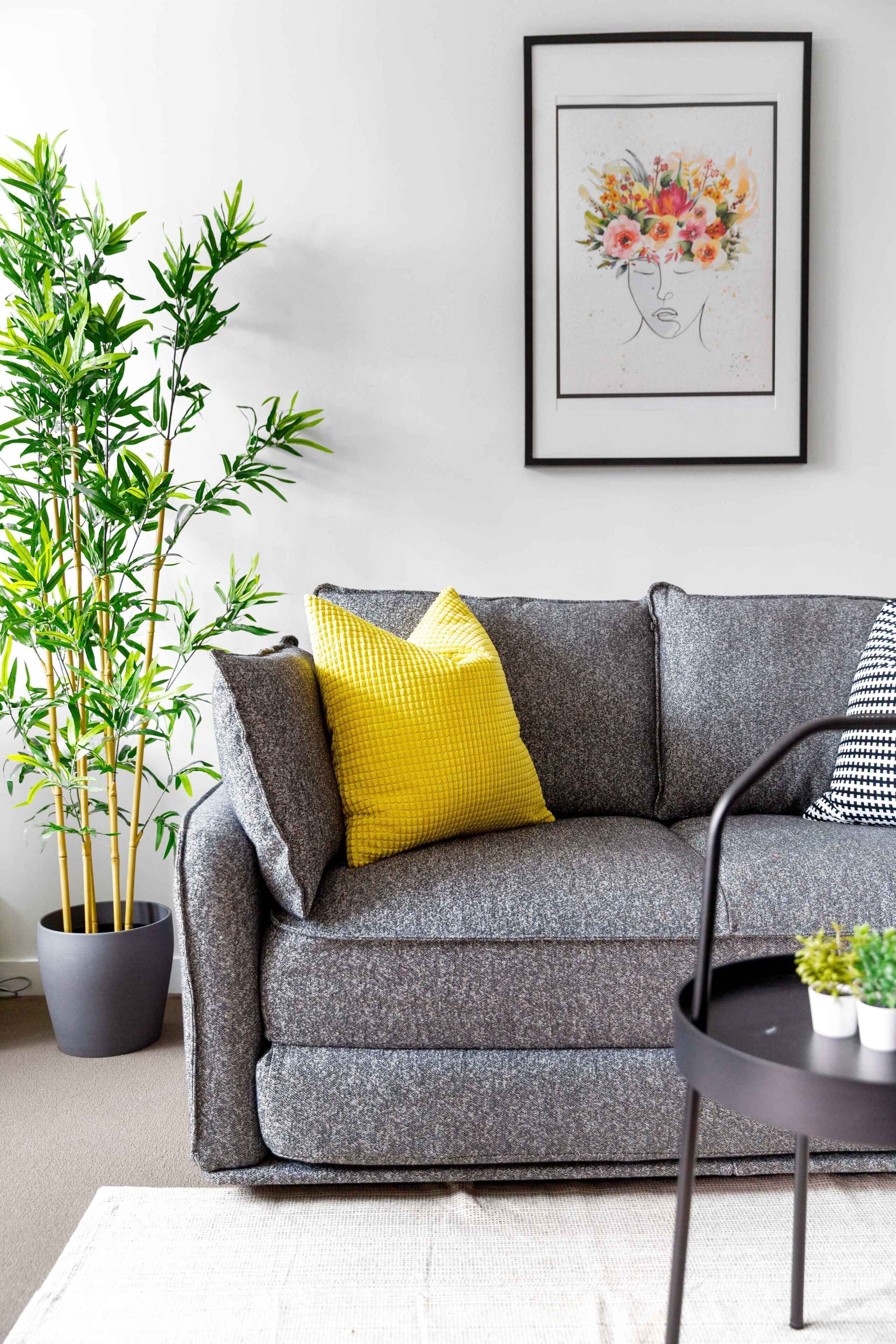 Un canapé gris agrémenté de coussins jaunes et noirs, une œuvre d’art florale encadrée accrochée au mur, et une plante en bambou placée à côté.