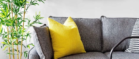 A grey sofa with yellow and black cushions, a framed floral artwork on the wall, and a bamboo plant beside it.