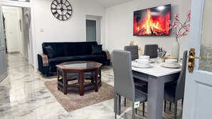 A dining area with a table set for four, a dark-colored sofa, a decorative clock, and a fireplace image on the wall. - Modern North London Retreat - 7 Bedrooms (London)